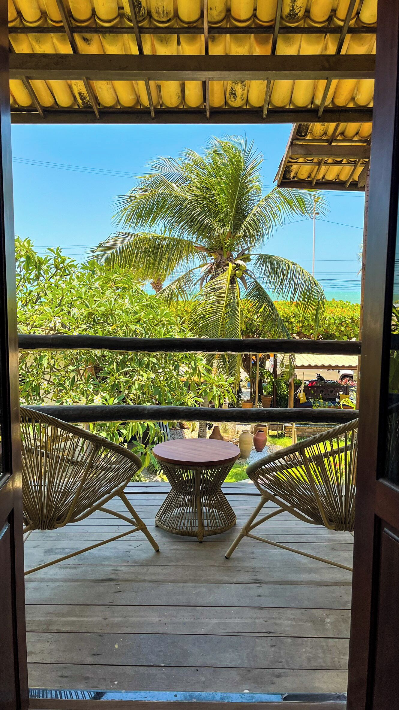 A wooden deck with a round table and two wicker chairs, overlooking a lush garden with a palm tree and various plants.