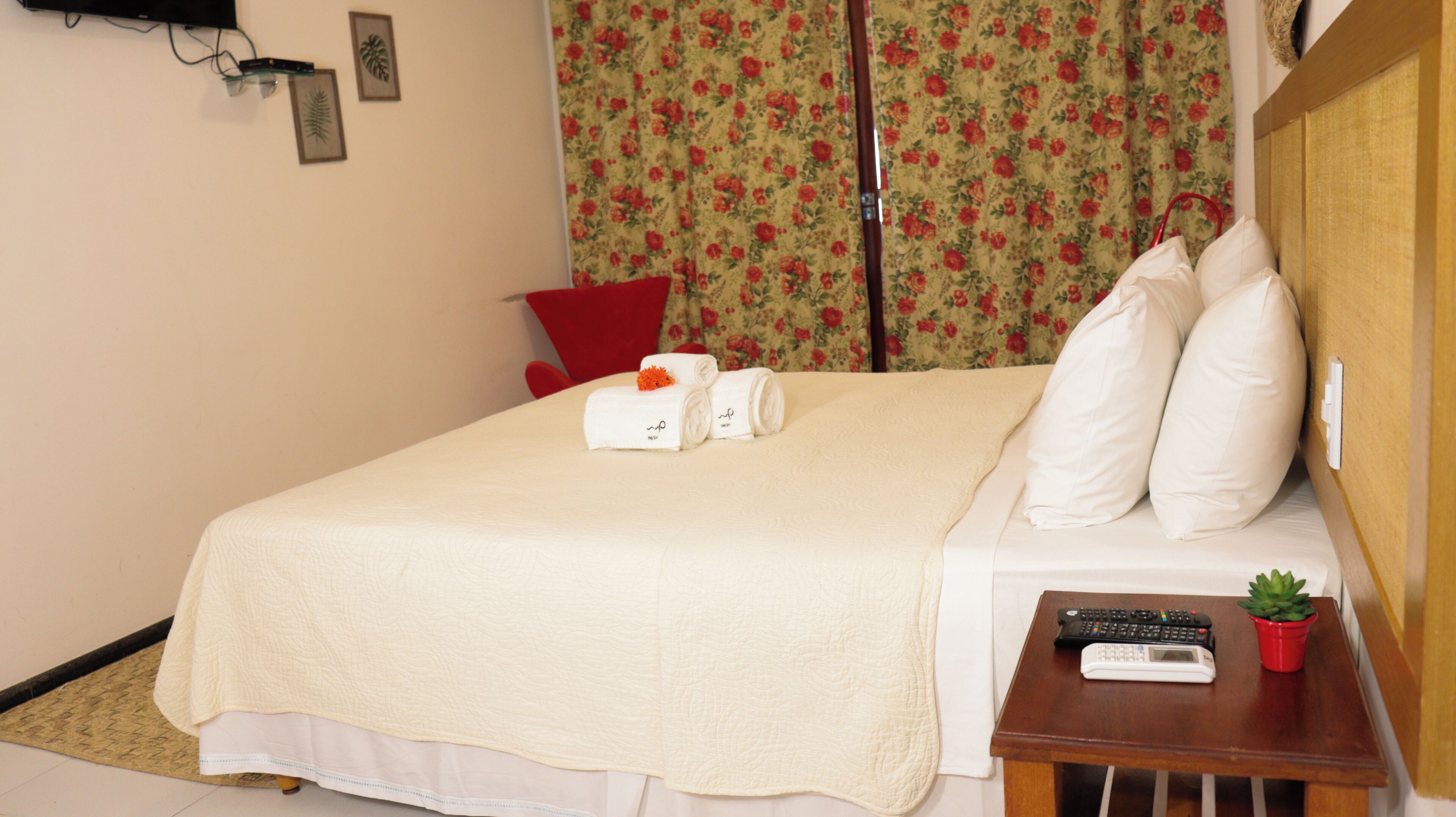 A hotel room with a bed, a red chair, a small table with a remote control, and a potted plant.