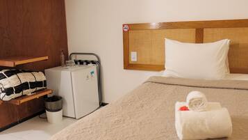 Een hotelkamer met een bed, een kleine koelkast, een prullenbak en een plank met een deken en glazen.
