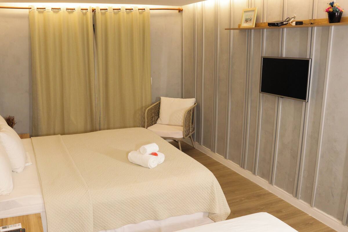 A hotel room with a bed, a chair, a TV, and a wall-mounted shelf with a picture frame and flowers.