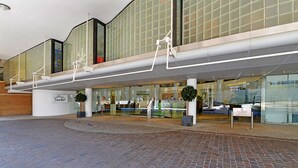 Un edificio moderno con fachada de vidrio, una zona de entrada cubierta y un patio adoquinado con ladrillos.