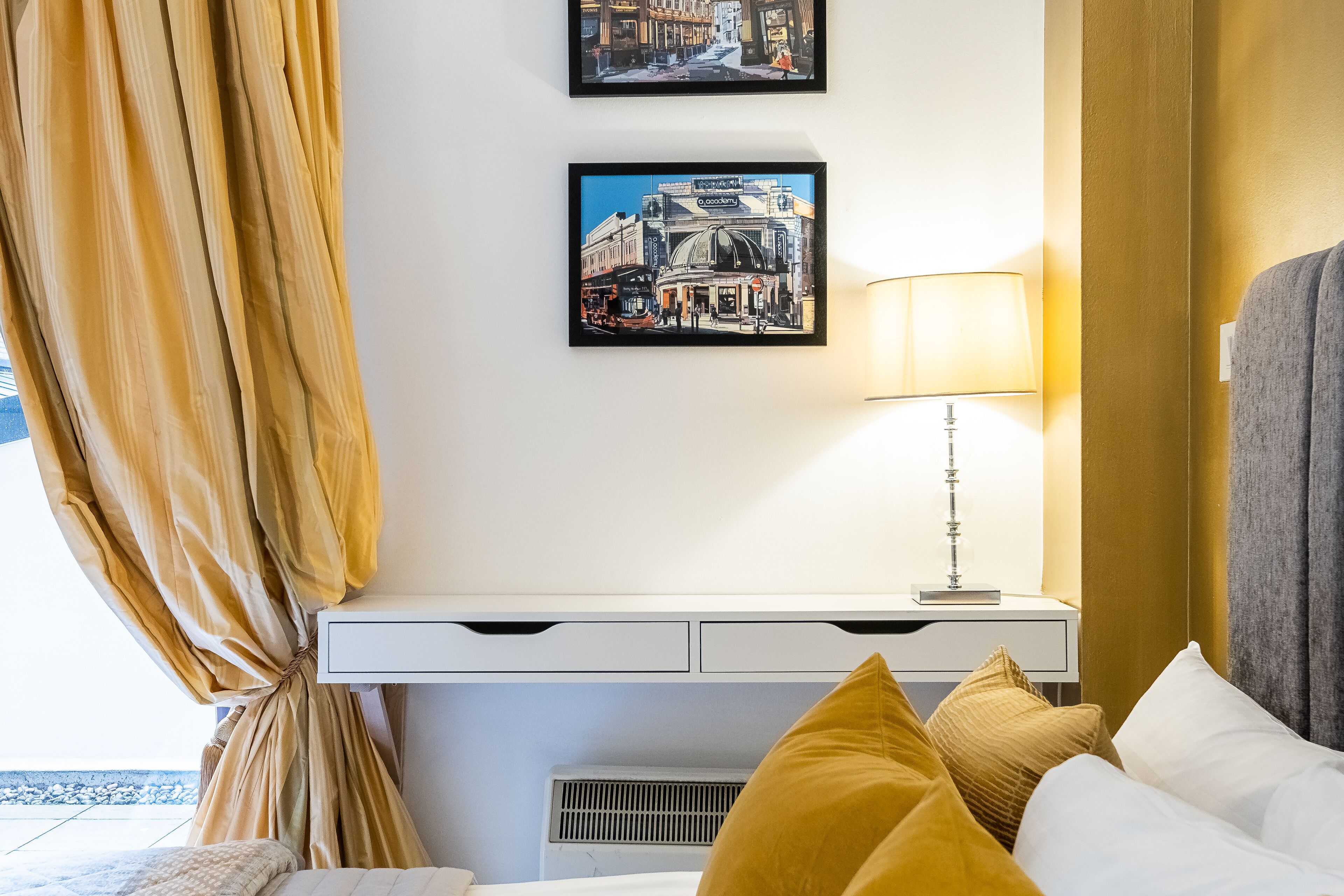 A hotel room with a bed, a bedside table with a lamp, a wall-mounted air conditioner, and two framed pictures.