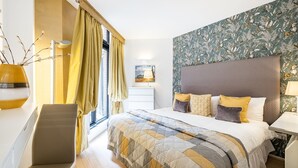 Un dormitorio con cama, escritorio, silla, ventana con cortinas, lámpara y pared con estampado floral.