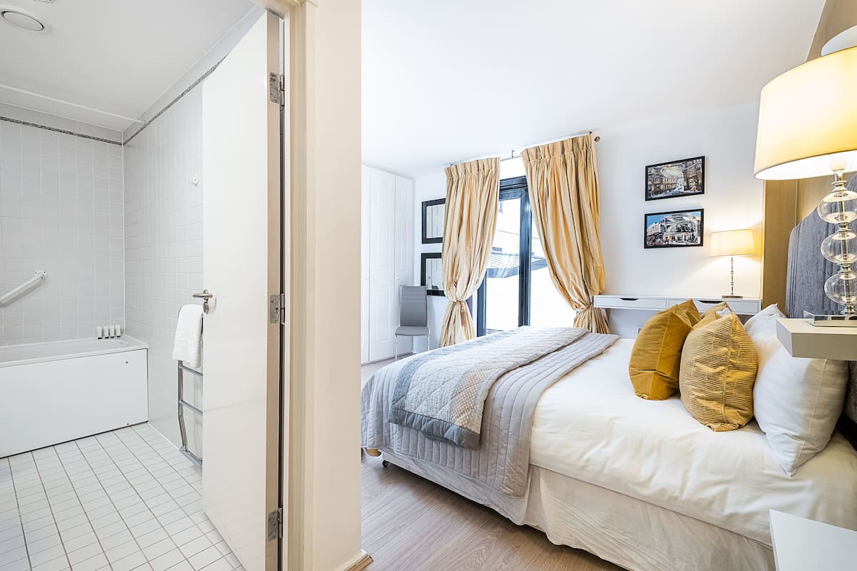 Ein Hotelzimmer mit einer weißen Badewanne, einem Bett mit gesteppter Decke und zwei Kissen, einem Nachttisch mit Lampe sowie einem Fenster mit Vorhängen.