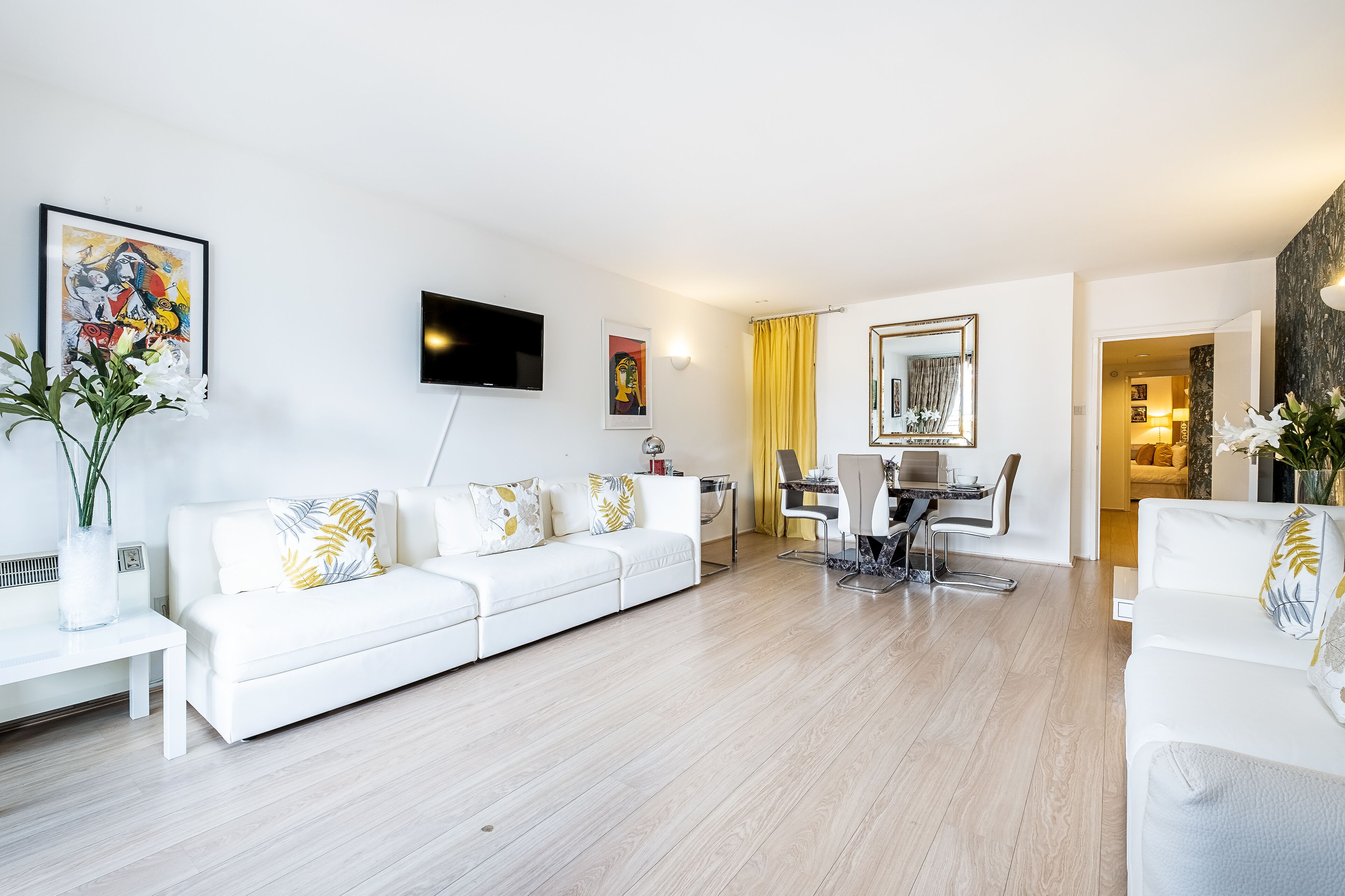 Una sala de estar moderna con un sofá blanco, una mesa de comedor con sillas, un televisor de pantalla plana y obras de arte en las paredes.
