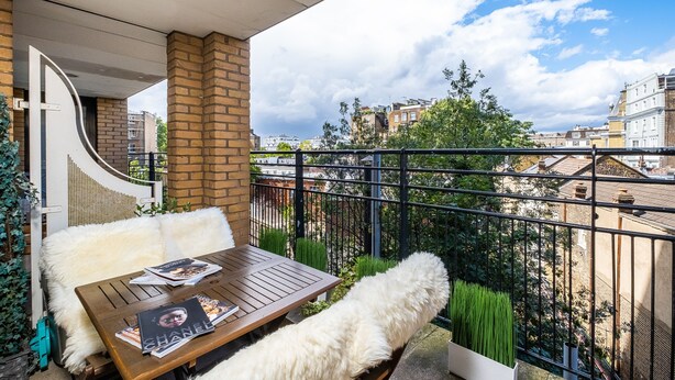Un balcón con una mesa de madera y sillas blancas y mullidas, con vistas a un paisaje urbano con edificios y zonas verdes.