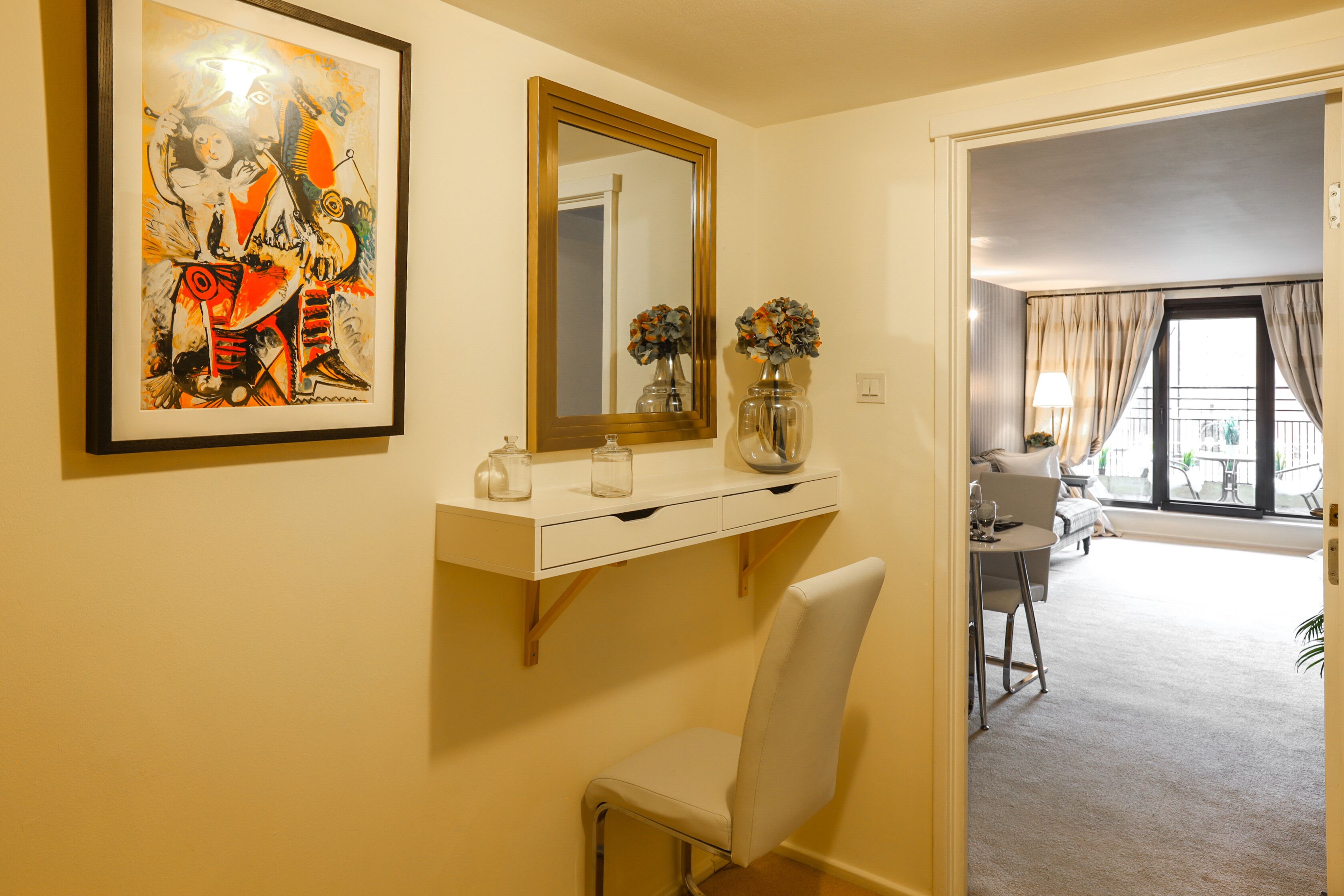 A hotel room with a white desk, a chair, a large mirror, and a framed abstract painting. There is a window with curtains and a view of the outside.