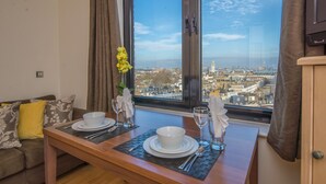 Un comedor con una mesa lista para dos personas, vista a un paisaje urbano a través de un ventanal grande, y un sofá con cojines.