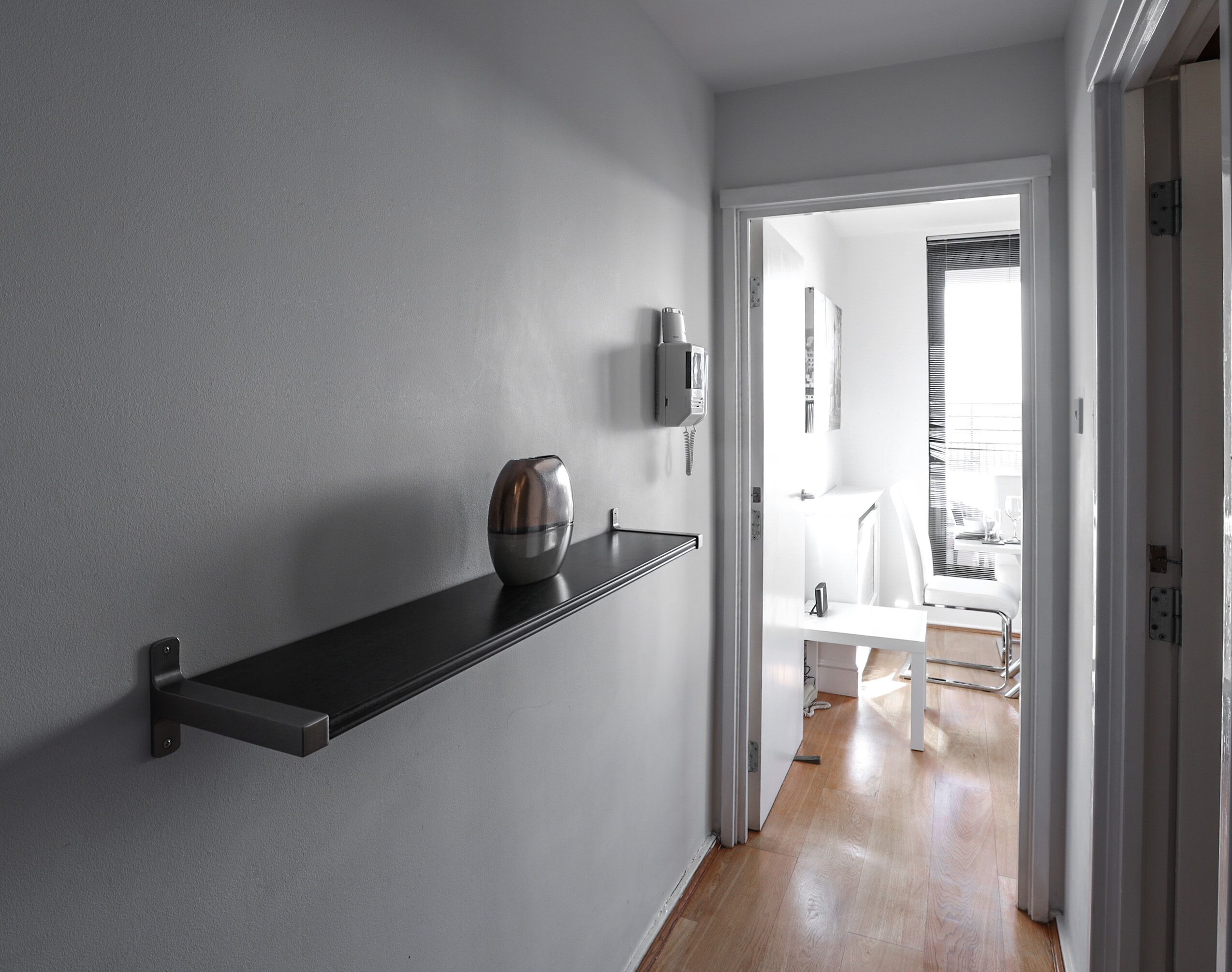 Un pasillo con un estante negro que sostiene un jarrón, un teléfono de pared y una puerta que da a una habitación con cama y ventana.