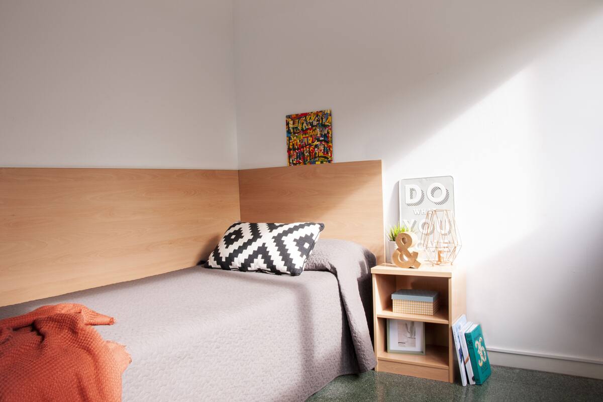 Une chambre à coucher bien aménagée, avec une tête de lit en bois, un lit agrémenté d’un coussin à motifs, une petite table de chevet avec une lampe et une œuvre d’art colorée accrochée au mur.