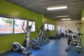 Ett gym med olika konditionsmaskiner, utrustning för styrketräning och fönster med utsikt över en byggnad med tegelstensfasad.