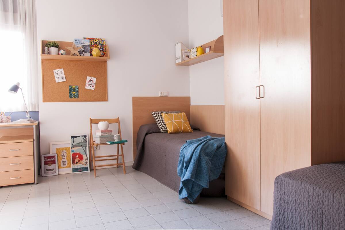 Une chambre à coucher comprenant un lit, un bureau, une chaise, une armoire et un tableau d’affichage en liège où sont accrochées des photos et des notes.