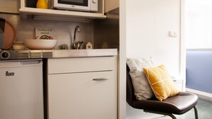 A compact kitchenette with a microwave, sink, and a small refrigerator. A chair with a cushion is visible next to the window. - Residencia Universitaria Resa Tomás Alfaro Fournier - Centro Adscrito a REAJ - Campus Accomodation (Vitoria-Gasteiz)