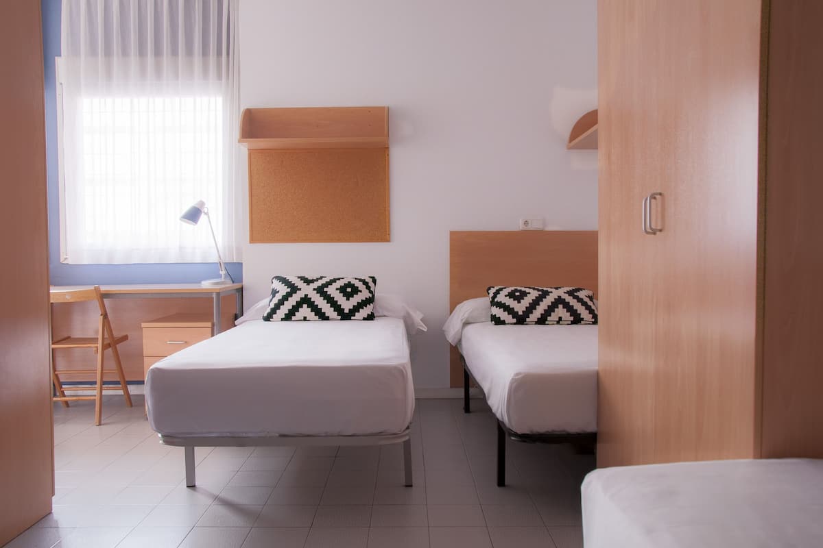 Une chambre d’hôtel avec deux lits simples, un bureau, une chaise et un tableau d’affichage en liège au mur.