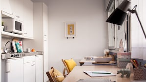 A modern kitchen with white cabinets, a microwave, and a dining area with a wooden table, chairs, and a bowl. - Residencia Universitaria Resa Tomás Alfaro Fournier - Centro Adscrito a REAJ - Campus Accomodation (Vitoria-Gasteiz)