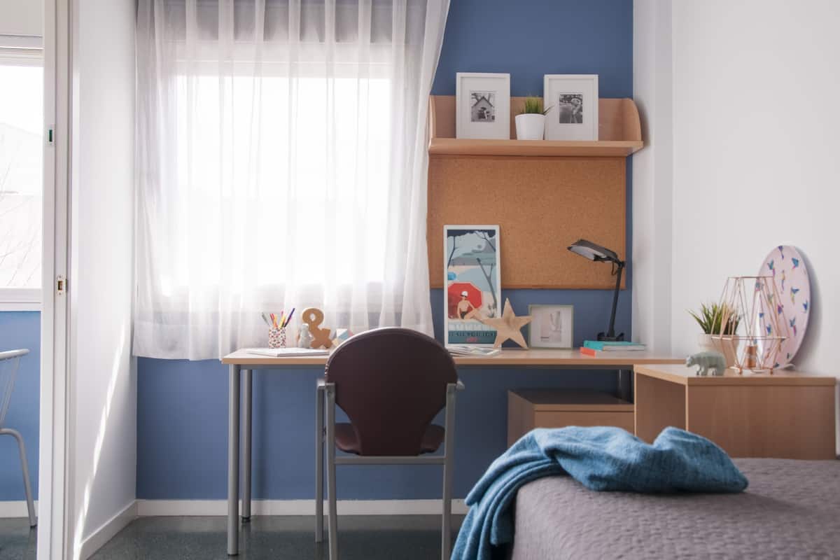 Une chambre à coucher avec un lit, un bureau, une chaise et une fenêtre ornée de rideaux légers.