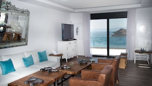 A modern living room with a white sofa, blue cushions, a wooden coffee table with decorative fish, a white cabinet, a flat-screen TV, and a large window with a view of the sea and a mountain. - Villa Dimitra by Elounda Island View Villas (Agios Nikolaos)