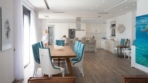 A modern kitchen with white cabinets, a wooden dining table with blue chairs, and a large painting of a seascape. - Villa Dimitra by Elounda Island View Villas (Agios Nikolaos)