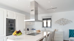 A modern kitchen with white cabinetry, a stainless steel oven, a central island with a sink, and a decorative wall piece. - Villa Dimitra by Elounda Island View Villas (Agios Nikolaos)