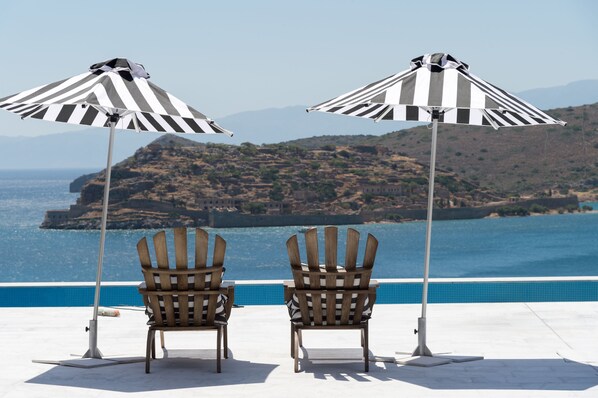 Two Adirondack chairs with striped umbrellas by a poolside, overlooking a coastal landscape with hills and the sea.