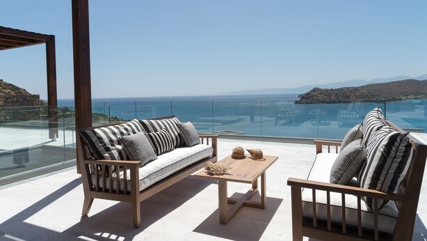 A modern outdoor seating area with a wooden sofa and armchairs, a coffee table, and a glass railing overlooking a coastal landscape. - Villa Dimitra by Elounda Island View Villas (Agios Nikolaos)