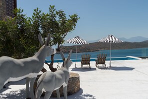 A poolside area with two Adirondack chairs and striped umbrellas, a statue of a white animal, and a view of a body of water and hills. - Villa Dimitra by Elounda Island View Villas (Agios Nikolaos)