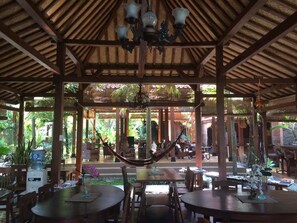 An outdoor dining area with wooden tables and chairs, a hammock, and a view of the surrounding greenery. - Bedhot Homestay (Sewon)