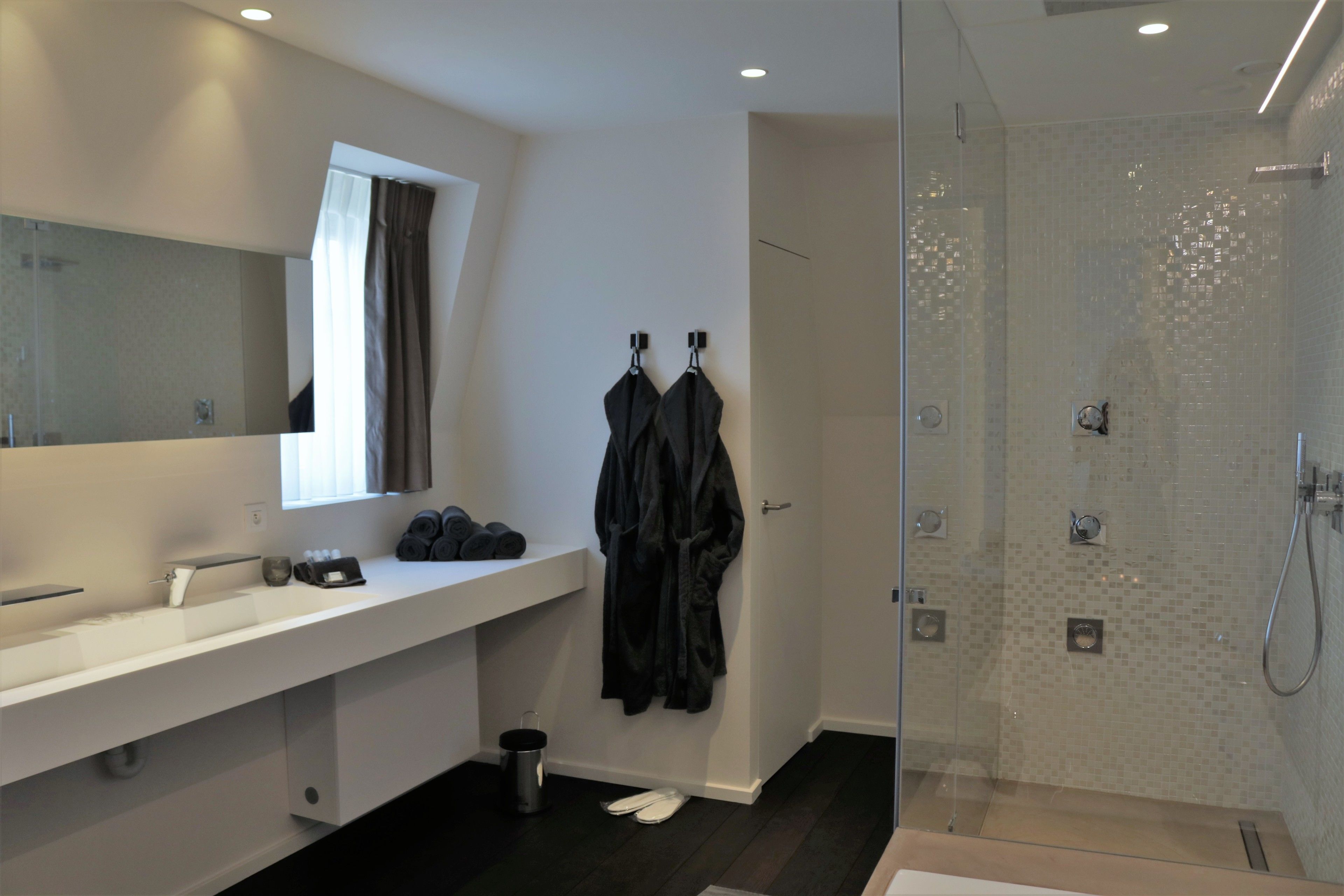 A modern bathroom with a large mirror, a white sink, a glass shower enclosure, and a robe hanging on hooks.