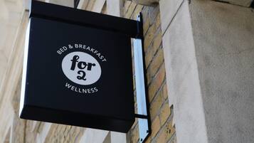 A black sign with white text reading "Bed & Breakfast for 2 Wellness" mounted on a brick wall.
