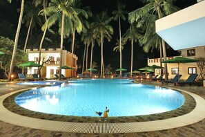 A swimming pool surrounded by lounge chairs and umbrellas, with palm trees and a building in the background.