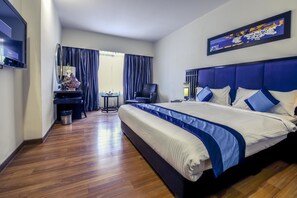 Ein Hotelzimmer mit einem großen Bett, einem Schreibtisch mit Stuhl, einem Fernseher und einem Fenster mit Vorhängen.