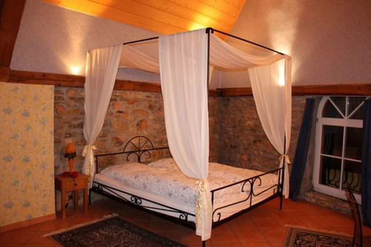 A four-poster bed with white drapes, a stone wall, and a window with curtains.