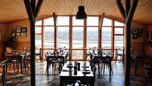 Een restaurant met grote ramen met uitzicht op het landschap. Tafels en stoelen staan er klaar voor een maaltijd.