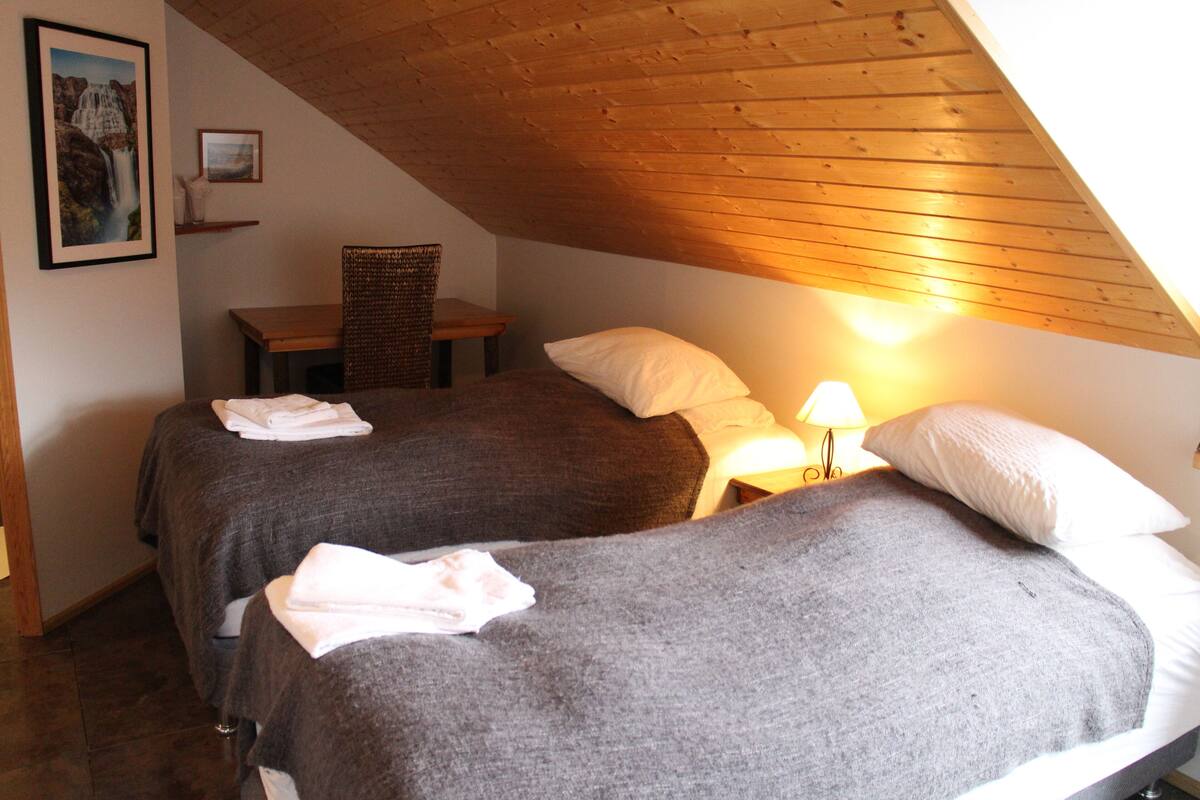 A bedroom with two beds, a wooden ceiling, and a nightstand with a lamp.