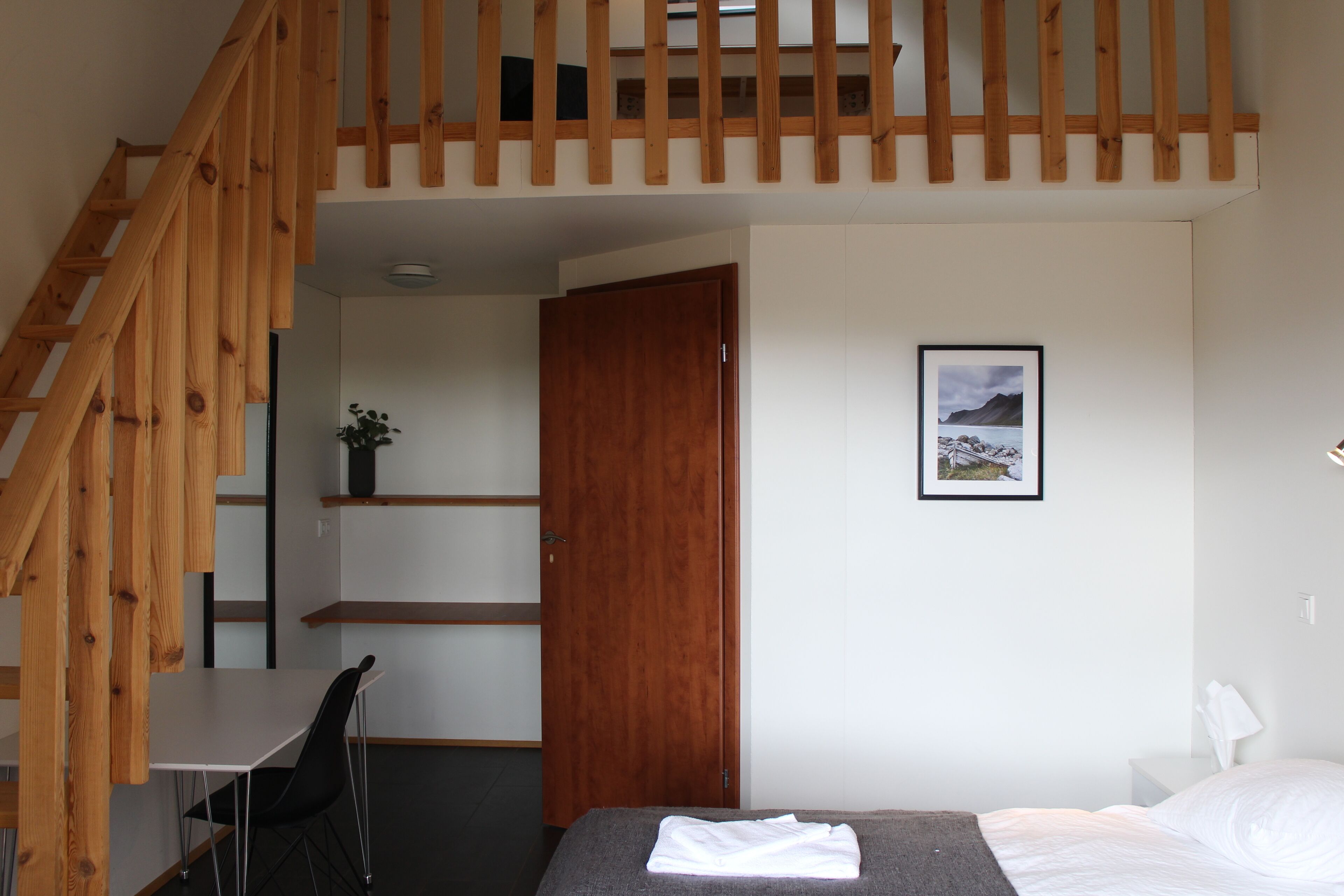 A bedroom with a wooden staircase, a bed with white bedding, a desk with a chair, and a framed picture on the wall.