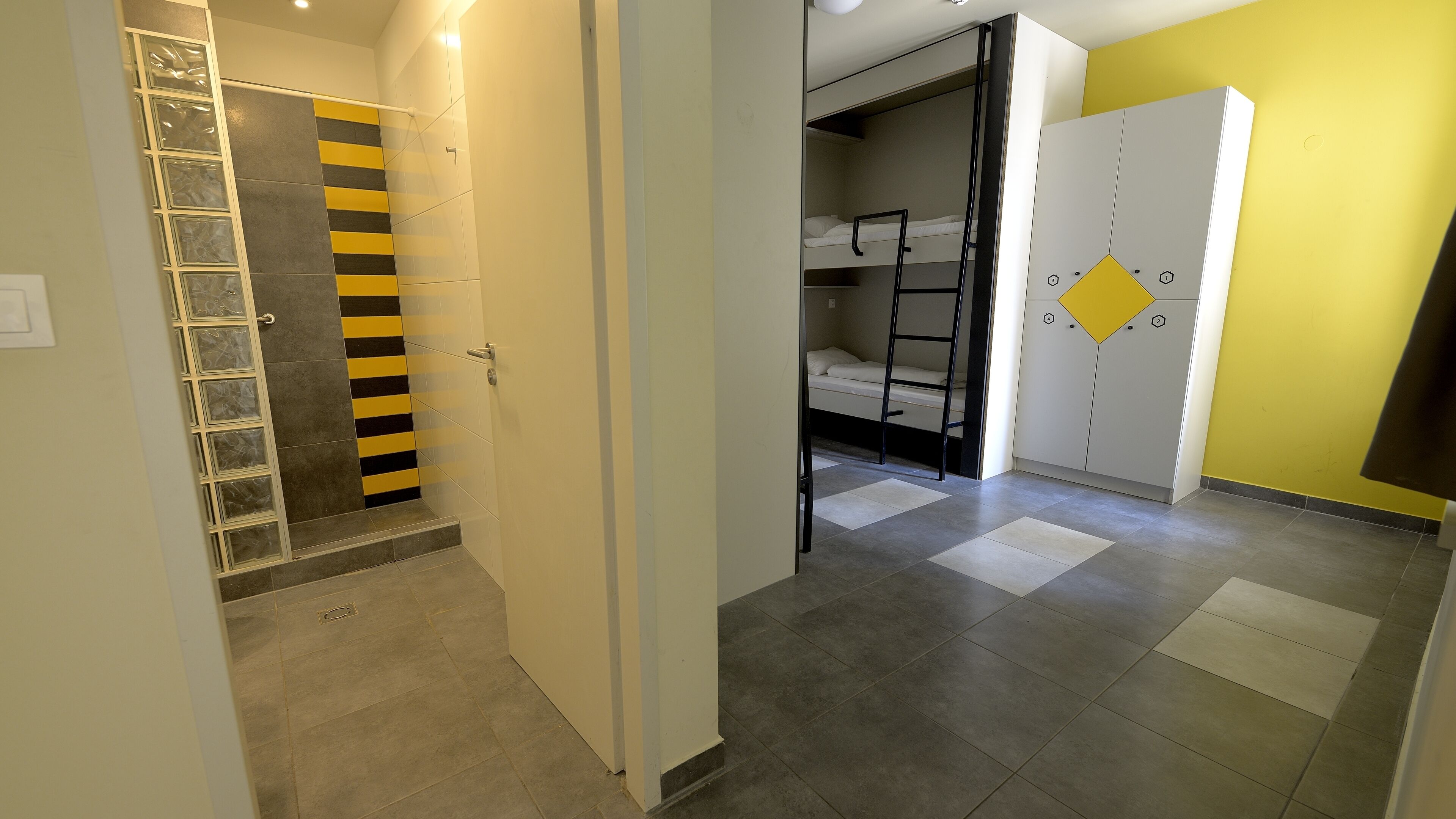 Um quarto com uma cama de solteiro, um guarda-roupa com detalhe amarelo e chão de azulejos.