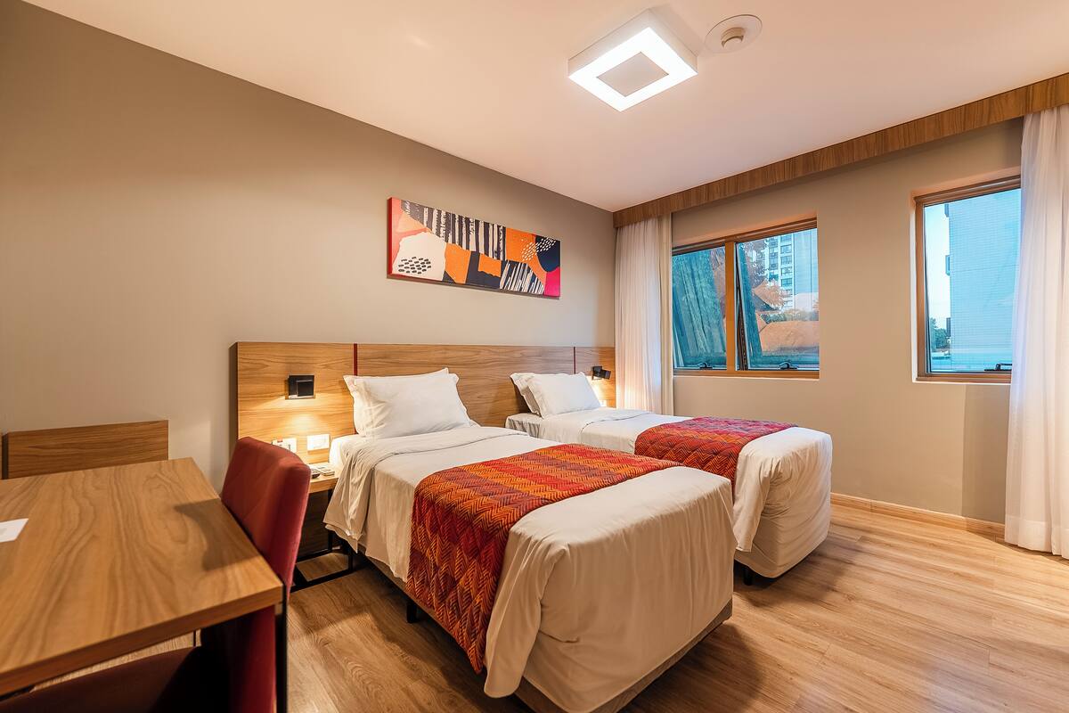 Une chambre d’hôtel avec deux lits, un bureau en bois, une chaise rouge, un tableau au mur et de grandes fenêtres avec des rideaux.
