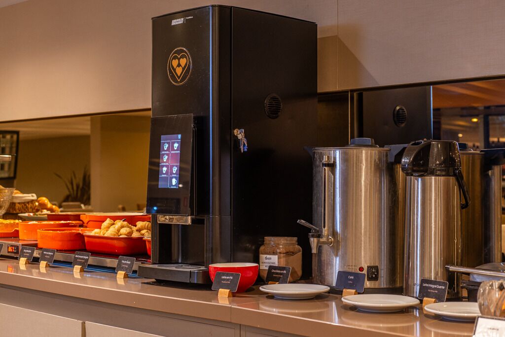 Uma área de buffet com máquina de café, panelas de aço inoxidável e recipientes de comida identificados.
