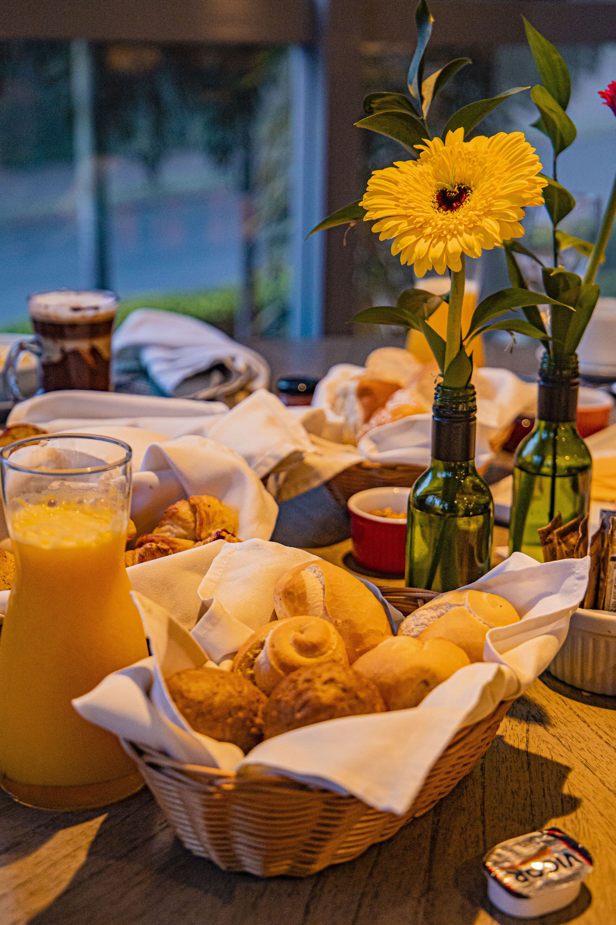 Una mesa de desayuno con pan, bollería, un vaso de zumo de naranja y dos botellas verdes.