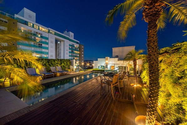 Un área de piscina con sillones reclinables, una terraza de madera y un edificio con varios pisos y balcones.