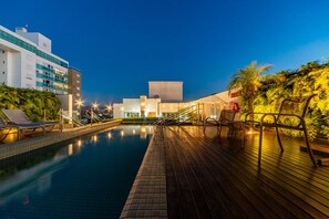 A swimming pool with a wooden deck, lounge chairs, and a building in the background. - Hotel Laghetto Stilo Higienópolis (Porto Alegre)