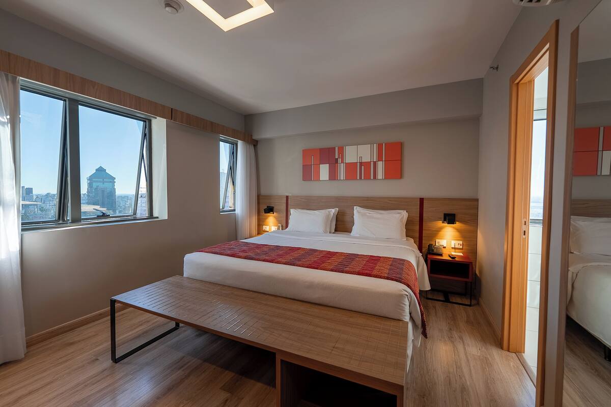 Une chambre d’hôtel moderne avec un grand lit, des tables de chevet avec des lampes, une vue sur la ville grâce à de grandes fenêtres et un panorama urbain visible à l’extérieur.