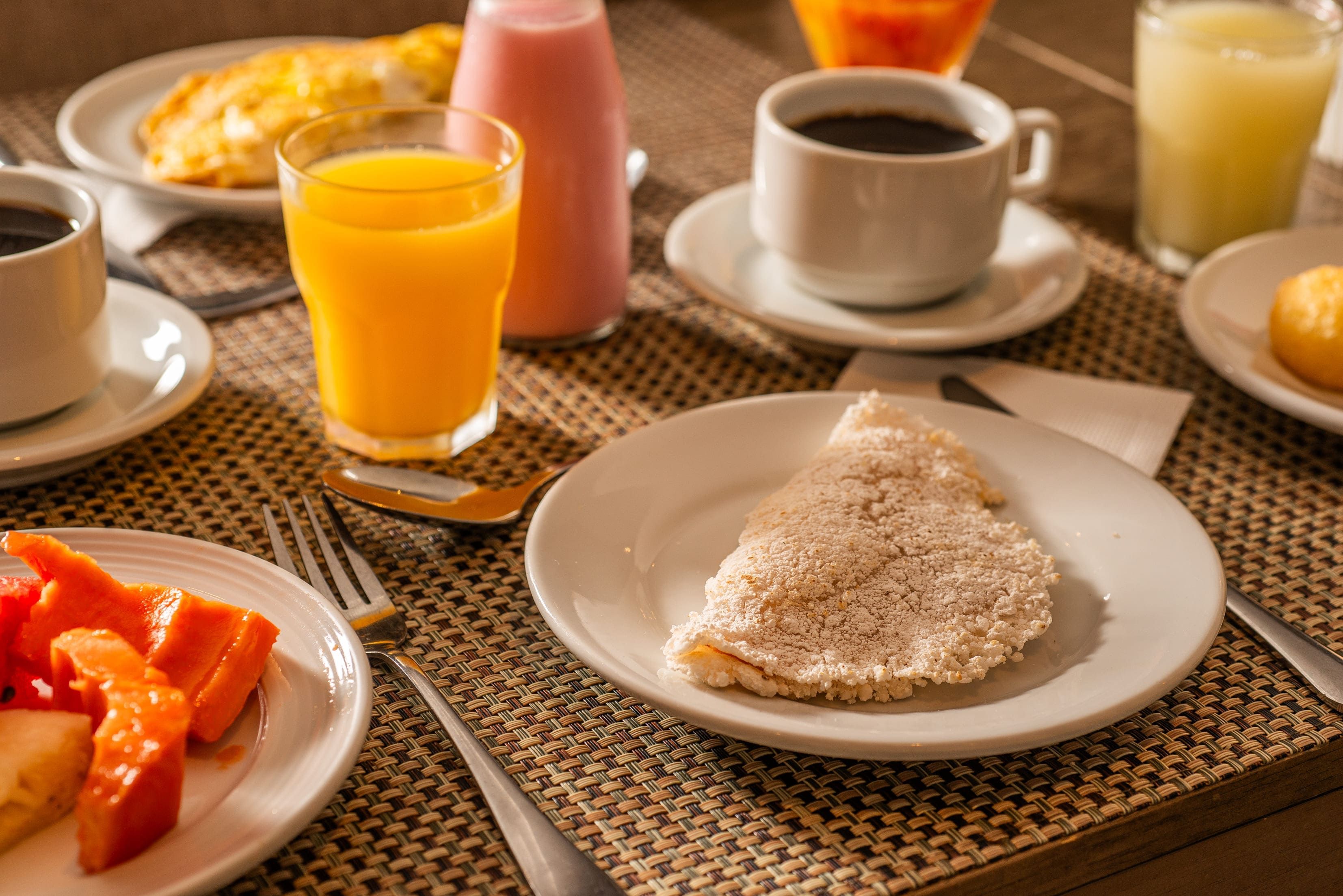 Mesa de café da manhã com pratos de comida, xícara de café e copo de suco de laranja.