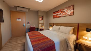 Quarto de hotel com cama, escrivaninha, ar condicionado, porta e obra de arte na parede.