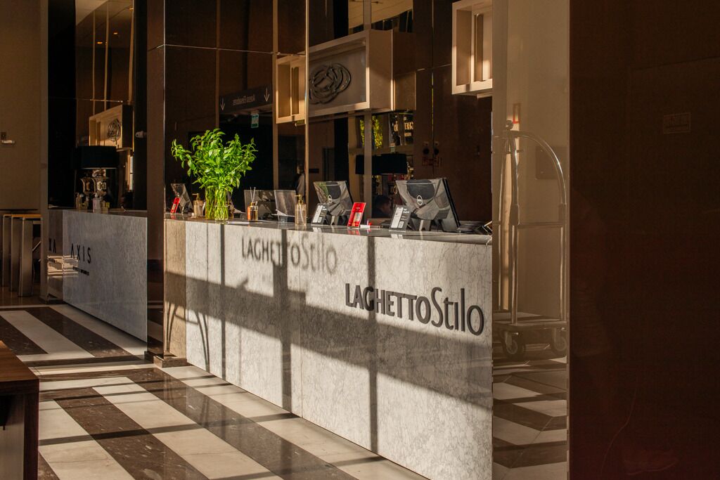 Uma mesa de recepção com balcão de check-in, uma planta em vaso e uma placa com a inscrição 'LAGHETTOStilo'.