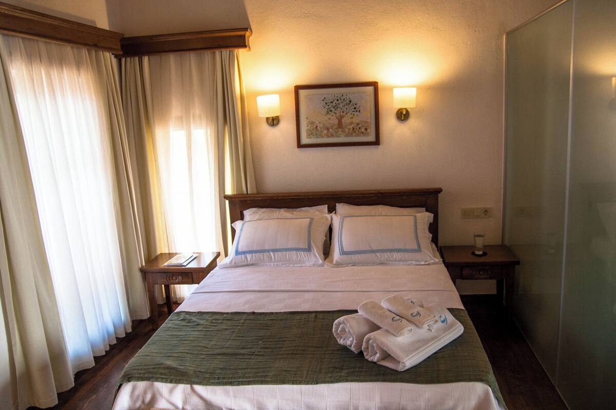 A hotel room with a bed, two bedside tables, a nightstand, a green throw blanket, and a folded towel on the bed.
