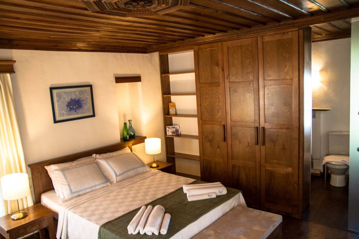 A hotel room with a bed, wooden wardrobe, a nightstand with a lamp, a framed picture on the wall, and a bathroom visible through an open door.