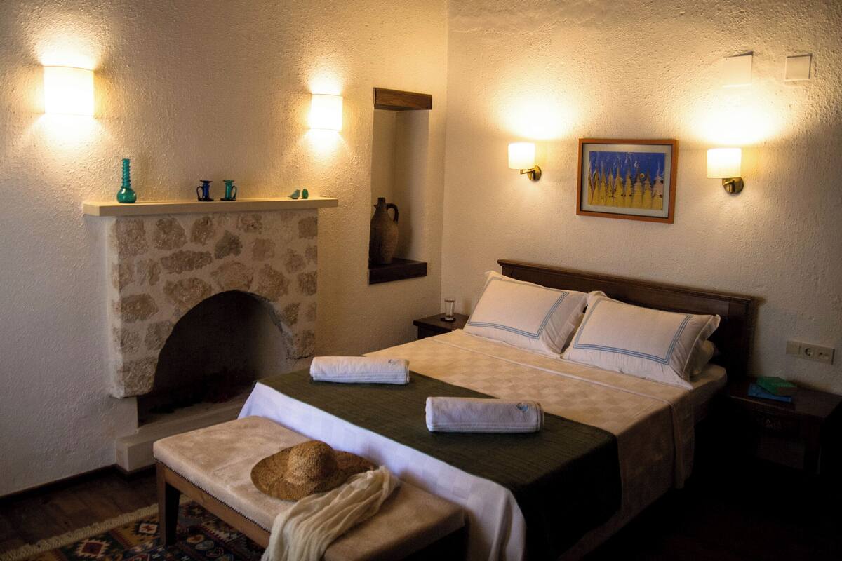 A bedroom with a stone fireplace, a bed with pillows, a bench with a hat, and wall-mounted lights.