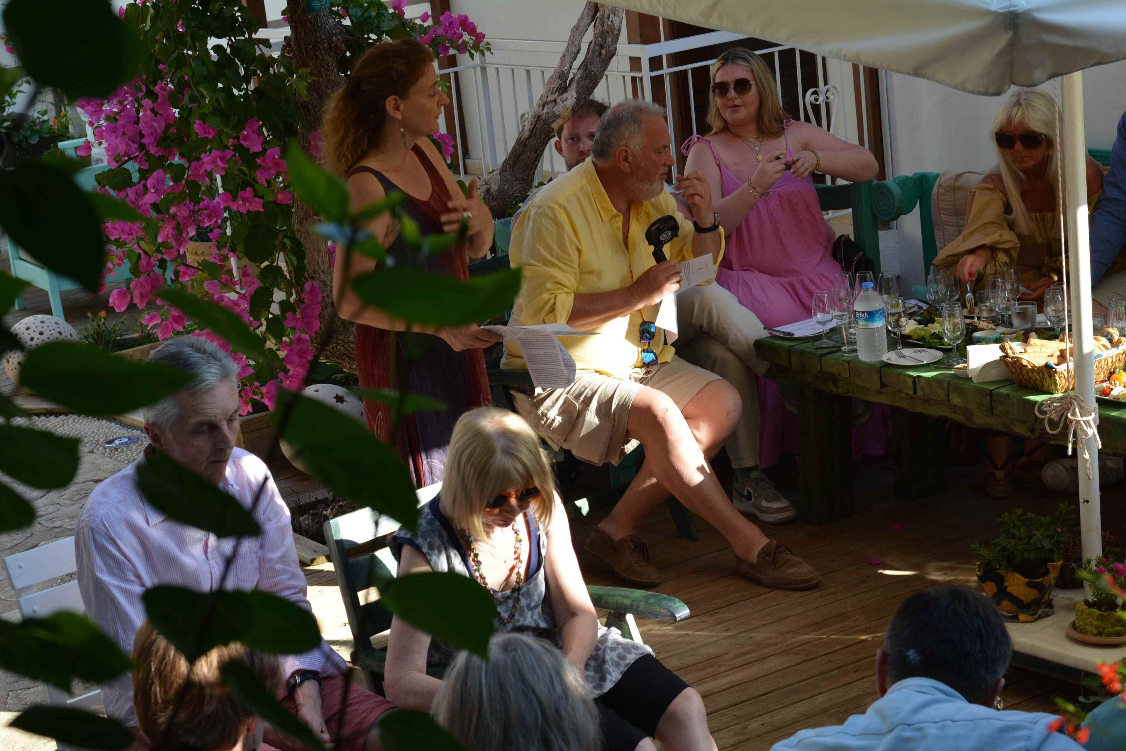 A group of people seated at an outdoor table with a tent overhead, surrounded by greenery and flowers.