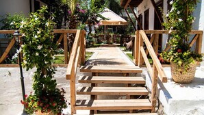 Una escalera de madera conduce a un espacio cubierto con techo de paja, rodeado de plantas en macetas y un edificio blanco al fondo.